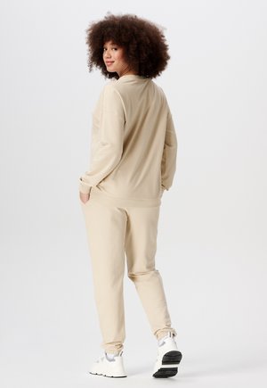 Beige Sweatshirt und passende Jogginghose, hergestellt aus weichem Stoff. Mit gerippten Bündchen, lockerer Passform und elastischem Taillenbund. Weiße Sneakers.