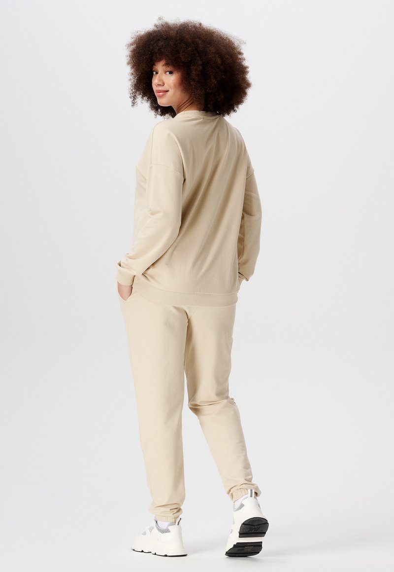 Beige sweatshirt en bijpassende joggers, gemaakt van zachte stof. Heeft geribbelde manchetten, een relaxte pasvorm en een elastische tailleband. Witte sneakers.