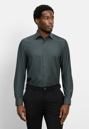 Mann trägt ein dunkelgrünes gemustertes Langarmhemd und schwarze Hose, steht vor einem schlichten weißen Hintergrund und schaut nach rechts.