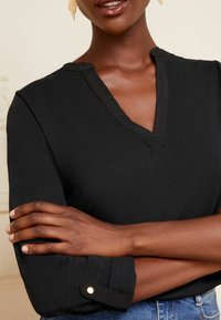 Haut noir à manches longues avec un décolleté en V, en tissu texturé, et détails de poignets boutonnés, associé à un jean bleu.