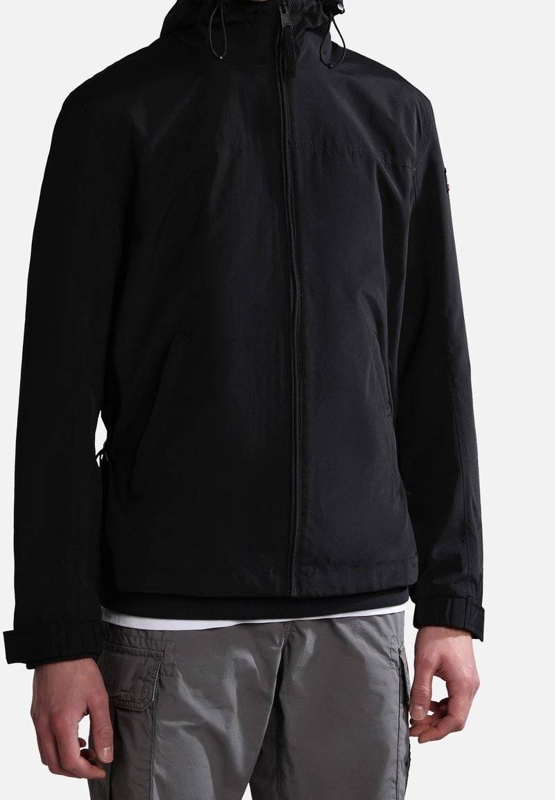 Giacca nera con zip e cappuccio, realizzata in un tessuto liscio e resistente all'acqua, completa di cordini regolabili e polsini elasticizzati. Abbinata a pantaloni grigi.