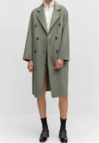 Manteau croisé en tissu vert doux, avec de grands boutons noirs, de larges revers et une coupe mi-longue. Porté sur un haut crème.