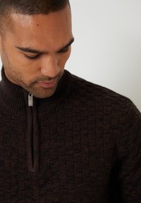 Brązowy sweter z zamkiem typu zip przy szyi, z teksturowanym wzorem w kwadraty, o ribbowanym kołnierzu i gładkim wykończeniu zamka. Ciepła, dziana tkanina odpowiednia na chłodniejsze dni.