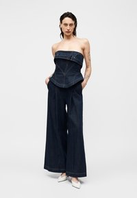 Kvinde iført mørkt denim stropløs korset-top med bukser med vide ben og hvide spidse sko, står med hænderne i lommerne.