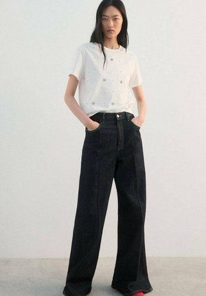 Cămașă albă cu mâneci scurte, cu modele florale brodate, asortată cu blugi din denim închis, cu talie înaltă și picioare largi. Accente roșii pe încălțăminte.