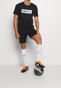Sort Nike F.C. t-shirt med hvidt logo, sorte shorts, hvide sokker og sorte fodboldstøvler med orange detaljer. Fodbold ved fødderne.