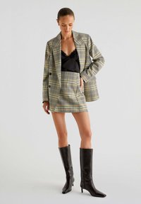 Rutet blazer og miniskørt i gul og sort nuancer, parret med en sort satin-camisole og knæhøje sorte læderstøvler.