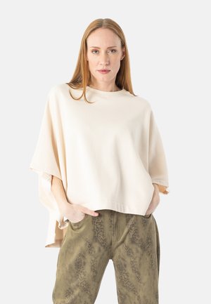 Kvinde med glat rødt hår iført en løs beige top og grønne bukser med mønster, stående med hænderne i lommerne foran en hvid baggrund.