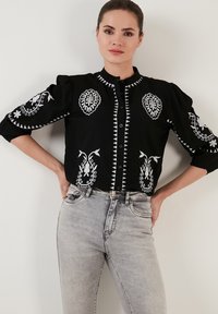 Schwarze Bluse mit weißer Stickerei, die florale und geometrische Muster aufweist, mit Puffärmeln und einer knöpfbaren Vorderseite. Getragen mit grauen Jeans.