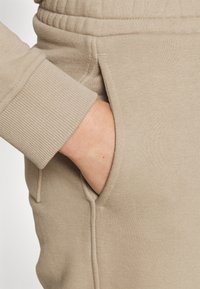 Ruka zasunutá v kapse béžových tepláků, manžeta rukávu žebrovaná a v odpovídající barvě materiálu.