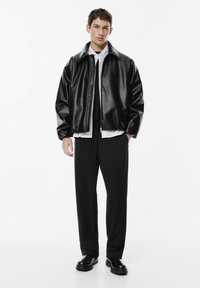 Jonge man die staat in een zwarte nep leren jas, wit shirt, zwarte wijdvallende broek en zwarte leren laarzen tegen een witte achtergrond.