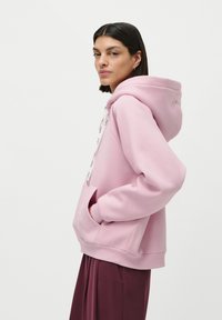 Femme aux cheveux bruns portant un sweat à capuche rose clair avec un logo sur les cordons et un pantalon bordeaux, posant de côté avec les mains dans les poches.