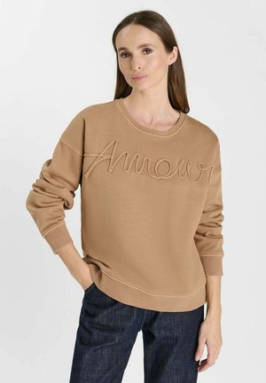 Donna con lunghi capelli castani che indossa una felpa beige con "Amour" ricamato e jeans blu scuro, in piedi con una mano in tasca.