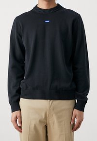 Mann in einem schwarzen, gerippten Rundhals-Pullover mit einem kleinen blauen "HUGO"-Logopatch und beigen Hosen, der vor einem einfarbigen Hintergrund steht.