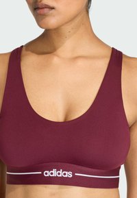 Burgundy sports bra made of stretchy fabric, featuring a wide underband with white "adidas" lettering and a scoop neckline.
