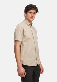 Beige overhemd met korte mouwen en een button-down kraag, subtiele textuur, en een klein logo op de borst, gecombineerd met donker geruite broeken.