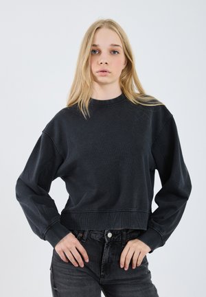 Svart cropped hettegenser med lange ermer, laget av mykt stoff. Har en rund halslinje og synlige sømdetaljer. Kombinert med mørke jeans.