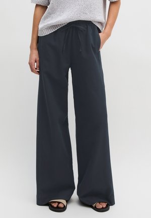 Osoba ubrana w luźne czarne spodnie z troczkiem, szary krótki sweter z dzianiny i czarno-beżowe klapki, stojąca z jedną ręką w kieszeni.