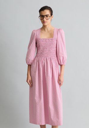 Kobieta ubrana w długą różową sukienkę w paski z bufiastymi rękawami do ćwiartki i kwadratowym dekoltem, stojąca na jednolitym jasnoszarym tle.