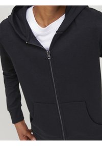 Zwarte rits-hoodie met een gladde textuur, kangaroezakken en een capuchon met trekkoord, gedragen over een wit T-shirt.