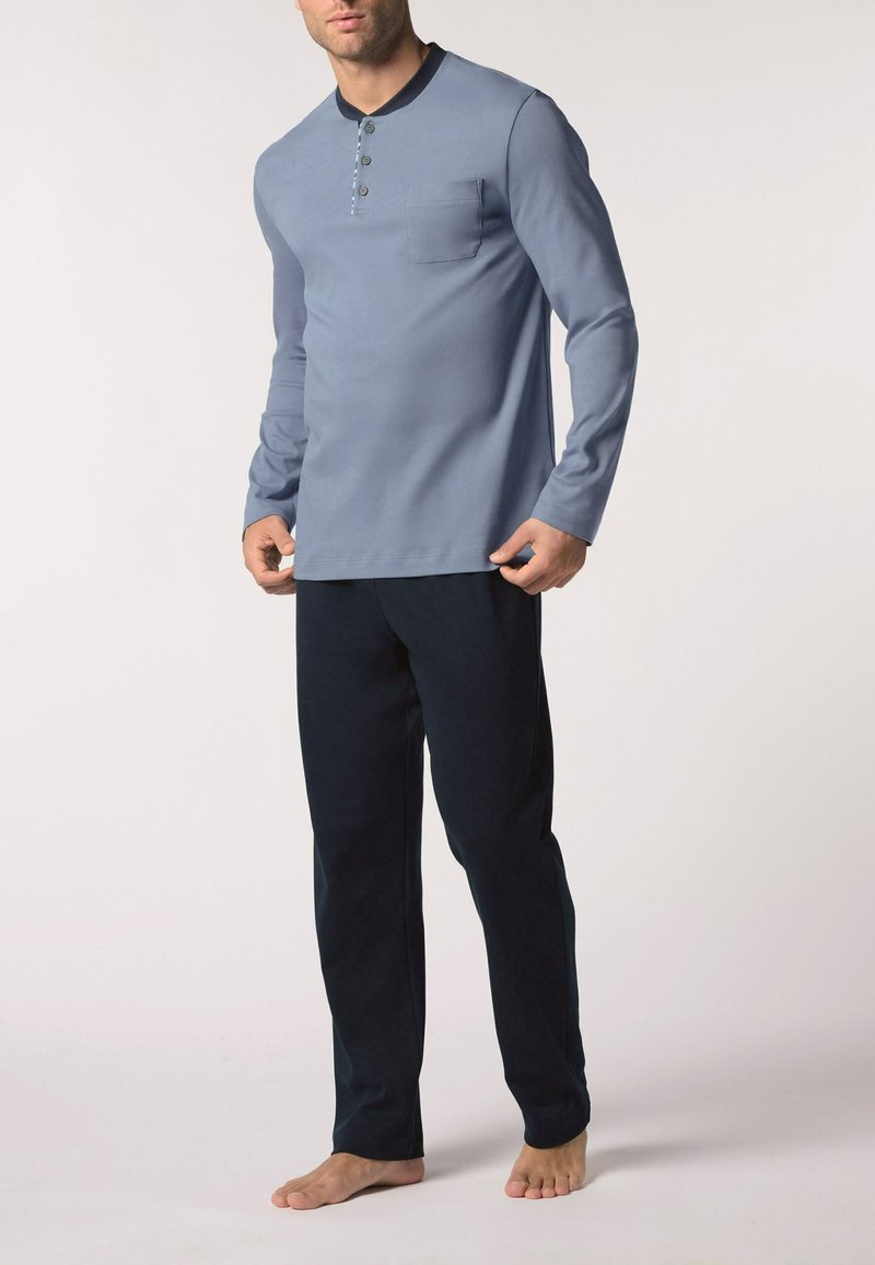 Maglietta henley a maniche lunghe di colore blu chiaro con tre bottoni e taschino sul petto, abbinata a pantaloni blu scuro. Tessuto morbido e liscio.