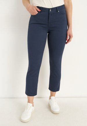 Kvinde iført marineblå korte bukser og hvide sneakers, stående med den ene hånd i lommen mod en ensfarvet baggrund.