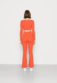 Conjunto de dois peças com padrão laranja, apresentando uma parte de cima cropped de mangas longas e calças flare, feito de material elástico. Usado com ténis brancos.