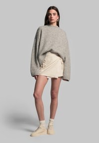 Hellgrauer, oversized Strickpullover mit weiten Ärmeln, kombiniert mit einem beigen Wickelrock mit Kordelzügen und cremefarbenen High-Top-Sneakern.