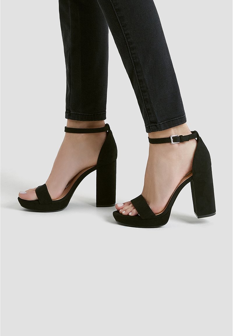 Pieds dans des sandales à talons blocs noirs avec des brides à la cheville et les orteils visibles, portant un pantalon noir ajusté sur un fond clair uni.