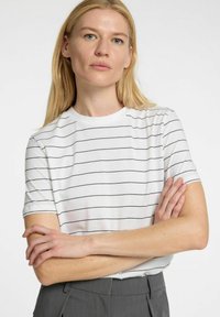 Vitt T-shirt med svarta horisontella ränder, korta ärmar, rund halsringning, slät textur, bärs med grå byxor, armarna korsade.