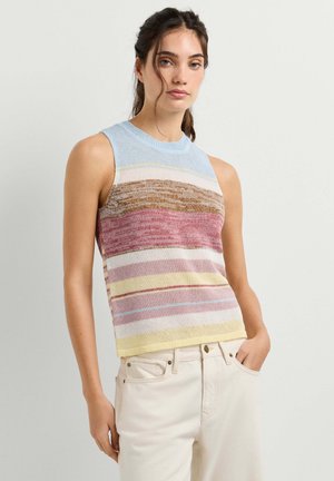 Jonge vrouw met een mouwloze, meerkleurige, gestreepte gebreide top en lichtbeige broek, staand met één hand in de zak tegen een effen achtergrond.