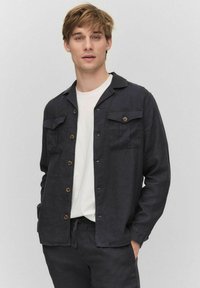 Donkerblauwe button-down shirt gemaakt van textiel stof, met een pointed collar, twee borstzakken en bruine knopen. Draagt over een wit shirt.