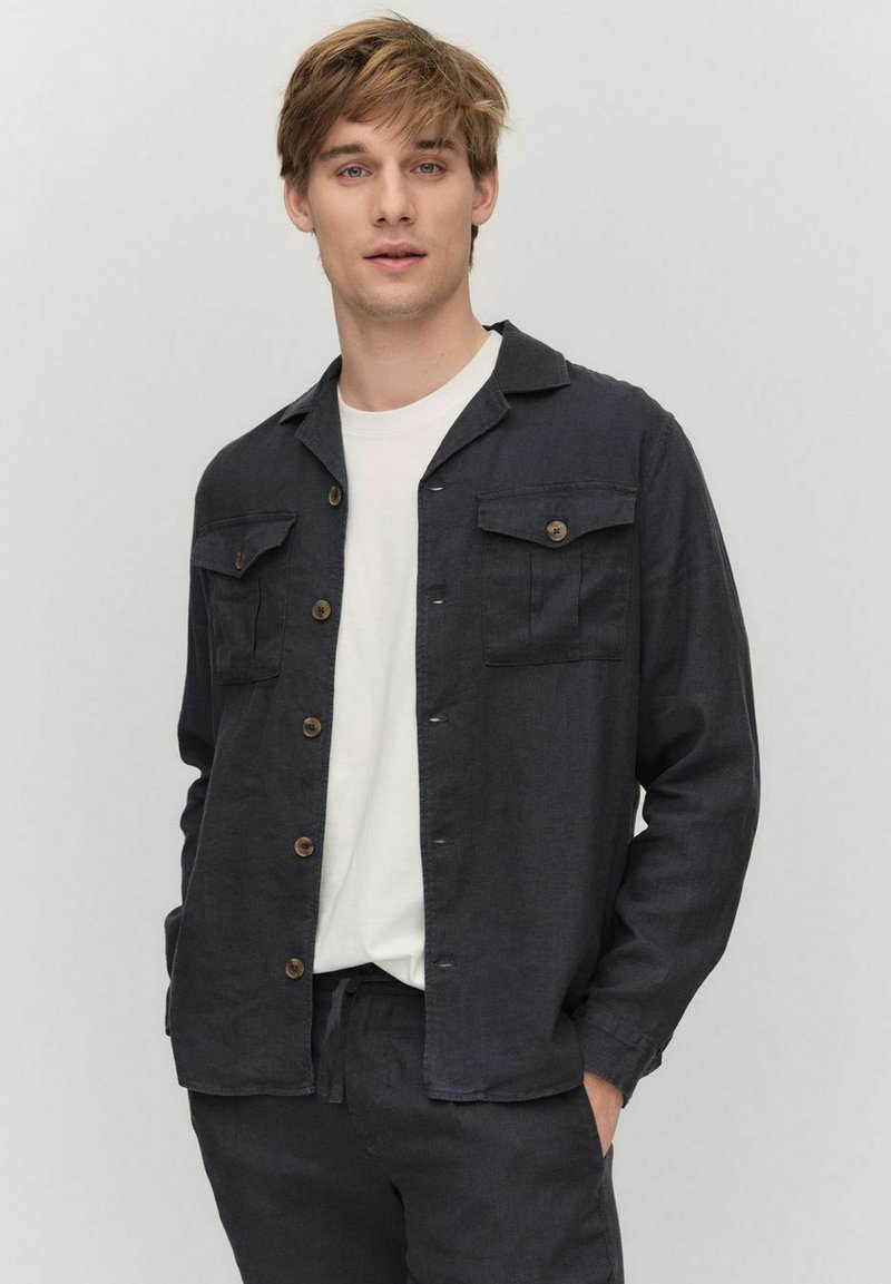 Donkerblauwe button-down shirt gemaakt van textiel stof, met een pointed collar, twee borstzakken en bruine knopen. Draagt over een wit shirt.