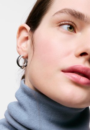 Zbliżenie osoby noszącej srebrną kolistą kolczyk i jasnoniebieski prążkowany golf, o jasnej karnacji i zielonych oczach.