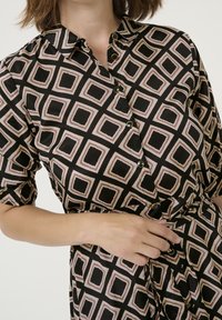 Chemise noire à manches courtes, avec un motif géométrique beige et noir, boutonnière à l'avant et un détail à taille cintrée.