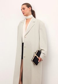 Per didelis šviesiai pilkas paltas su lygiu paviršiumi ir penkiais mygtukais, derinamas su juodu rankinuku su auksiniu užsegimu ir baltu šaliku.