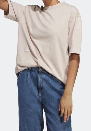 Persoon draagt een loszittend licht beige T-shirt met korte mouwen, wit opschrift en blauwe spijkerbroek, tegen een effen achtergrond.