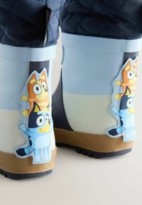 Las botas de lluvia de goma en azul y blanco tienen una suela marrón. Decoradas con pegatinas de personajes de dibujos animados en azul y naranja en los lados.