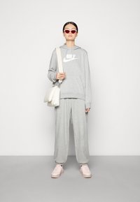 Grijze hoodie met wit logo, bijpassende sweatpants en lichtroze sneakers, aangevuld met een witte tas en rode zonnebril.