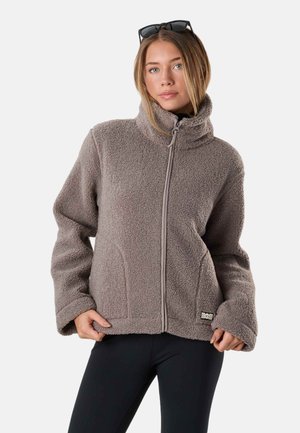 Kvinde iført grå fleecejakke med lynlås og sorte leggings, med solbriller hvilende på hovedet, stående mod en ensfarvet baggrund.