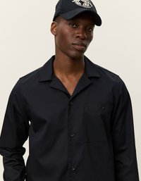 Camisa negra de botones con un bolsillo, cuello corto y mangas largas. Lleva puesta una gorra negra a juego con un logo. Textura suave.
