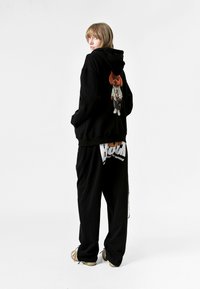 Sudadera negra con un gráfico de un oso en la espalda, combinada con pantalones de chándal negros que tienen letras blancas. Tejido casual, corte holgado.