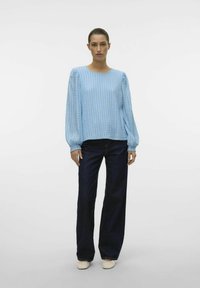 Heledsinine tekstuuriga pikkade varrukatega pluus ümmarguse kaelusega, koos tumehalli laia jalgadega teksapüksiga ja heledate slip-on kingadega.