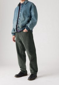 Chaqueta de mezclilla con un lavado azul claro, puños acanalados; pantalones de pana verde; zapatos negros de cordones. Fondo neutro.