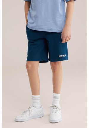 Gyermek tengerészkék rövidnadrágban, amelyen a bal lábszáron a "BAD BOYS" felirat olvasható, világoskék ingben, fehér zokniban és fehér tornacipőben áll.