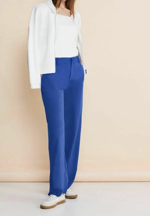 Mujer con chaqueta blanca sobre camiseta blanca, pantalones holgados azul real y zapatillas blancas, de pie contra un fondo beige.