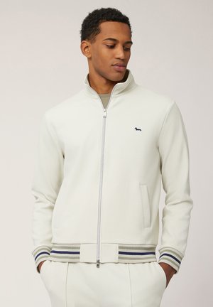 Veste à zip blanc avec des poignets côtelés, présentant des accents rayés gris et bleu, des poches latérales et un petit logo sur la poitrine.
