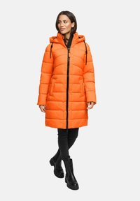 Mujer vestida con un abrigo de plumas largo y naranja con capucha, pantalones negros y botas negras hasta el tobillo, de pie contra un fondo liso.