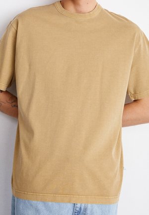 Person iført en ensfarvet beige T-shirt med korte ærmer og lyseblå jeans, stående med hænderne bag ryggen mod en hvid baggrund.