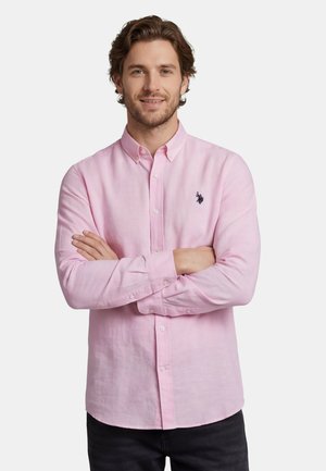 Mann mit kurzen braunen Haaren, der ein hellrosa Hemd mit Knöpfen und kleinem dunklem Logo trägt, steht mit verschränkten Armen vor einem weißen Hintergrund.